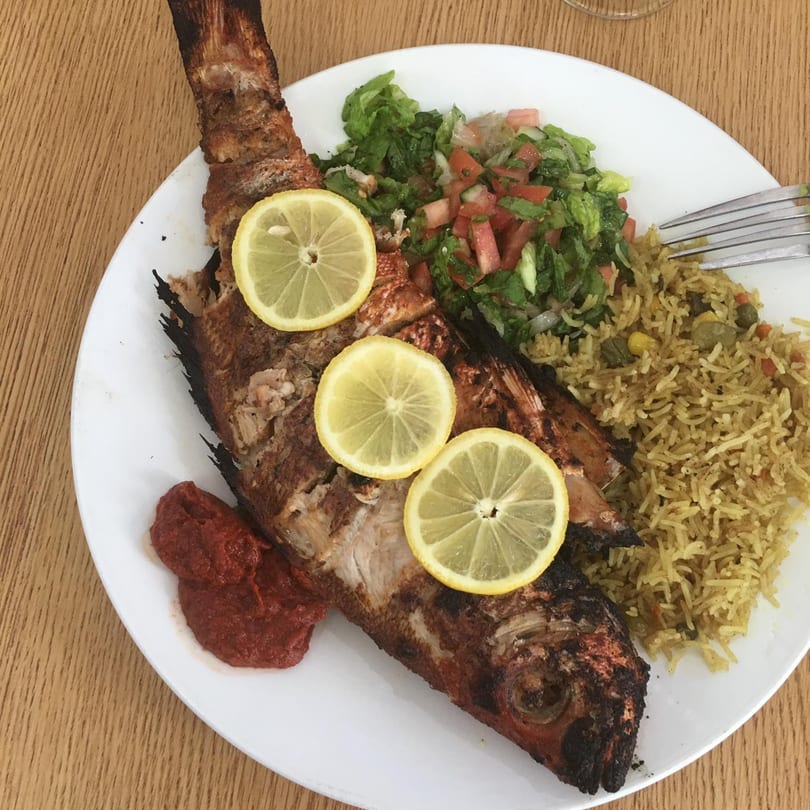 Whole grilled fish with lemon slices, rice, salad, and red sauce on a white plate.