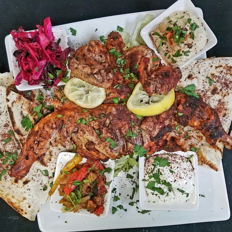 A large platter of grilled chicken with lemon, pita bread, pickled cabbage, hummus, and roasted vegetables.