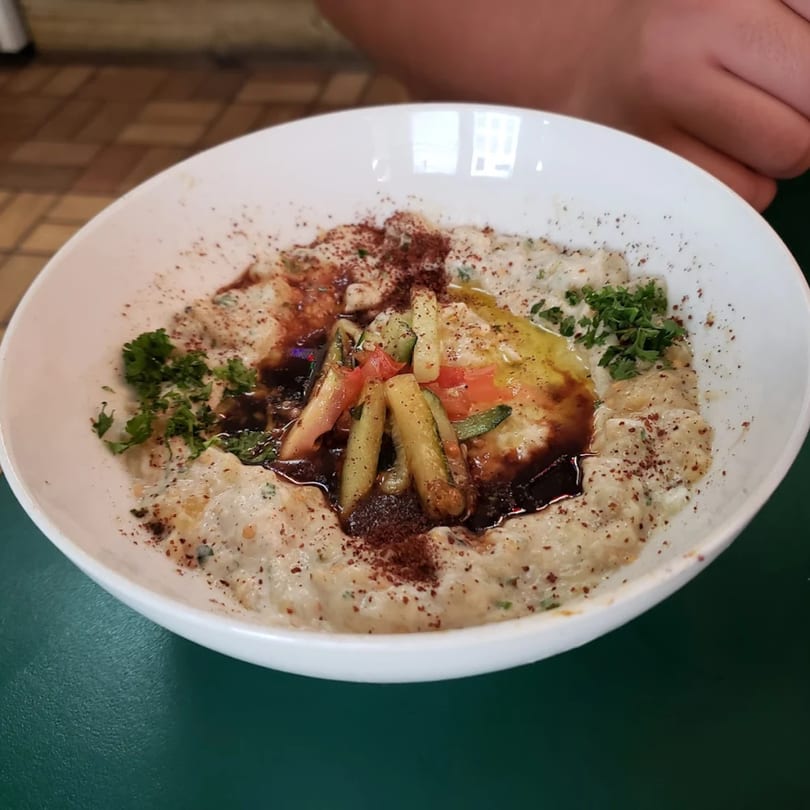 A white bowl of creamy baba ghanoush with olive oil, sumac, parsley, and pickled vegetables.