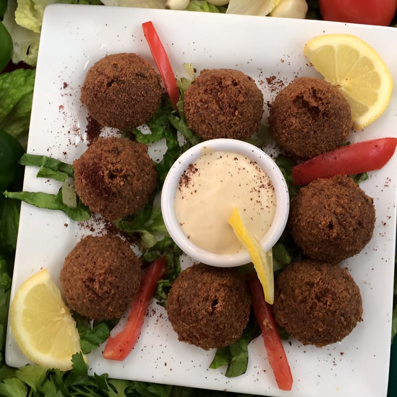 Nine falafel balls with a side of tahini sauce, lemon wedges, and tomato slices on a white plate.